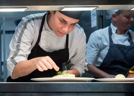 A chef preparing a plate of food in a kitchen with another chef stood behind them.