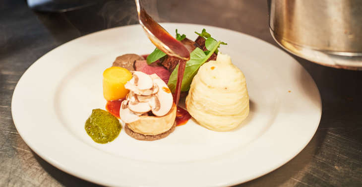 A chef plating up a dish in the kitchen, finishing it with a gravy topping.