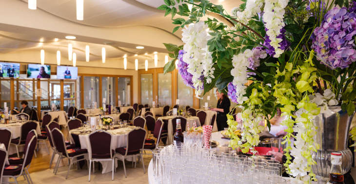 A bar with glasses and bottles of Pimm's and flowers on it in the foreground with tables set for dinner and staff in the background.