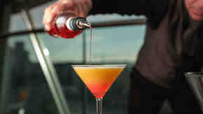 A person pouring a red liquid into a glass of an orange cocktail.