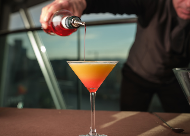 A person pouring a red liquid into a glass of an orange cocktail.