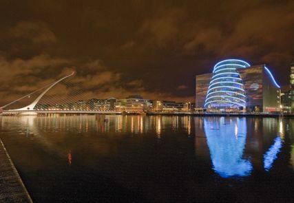 A night time shot of the Convention Centre Dublin lit up with blue lights and a view of the waterfront.