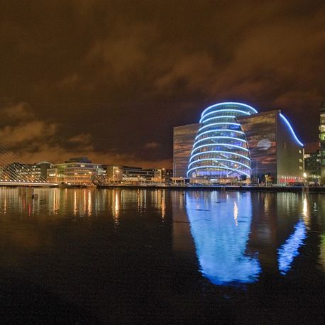 A night time shot of the Convention Centre Dublin lit up with blue lights and a view of the waterfront.