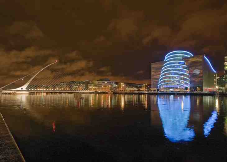 A night time shot of the Convention Centre Dublin lit up with blue lights and a view of the waterfront.