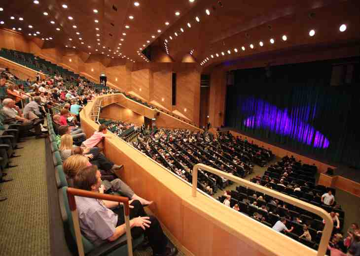 Crowds seated in rows of seats on a balcony and floor level in a theatre looking towards a stage.