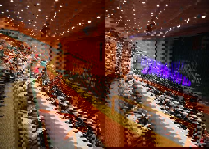 Crowds seated in rows of seats on a balcony and floor level in a theatre looking towards a stage.