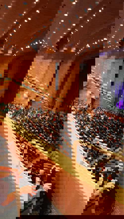 Crowds seated in rows of seats on a balcony and floor level in a theatre looking towards a stage.