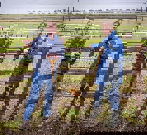 Two men stood with shovels in an empty field with the racecourse behind them.
