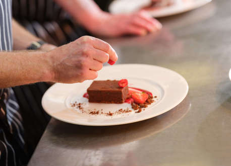 A hand preparing a chocolate cake with sliced strawberries on a white plate.