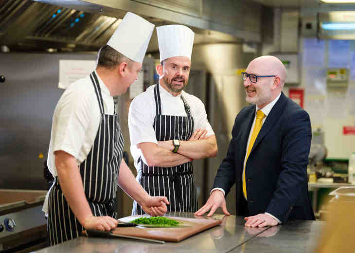 A man in a suit talking to 2 chefs in a kitchen.