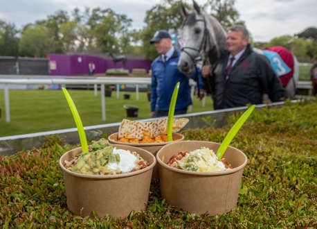 3 bowls of food resting on a hedge whilst 2 men lead a horse in a background at a racecourse.