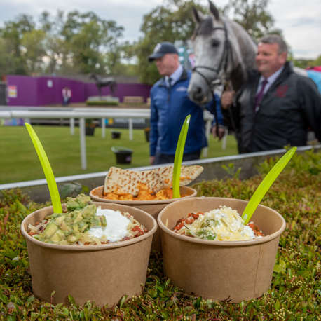 3 bowls of food resting on a hedge whilst 2 men lead a horse in a background at a racecourse.