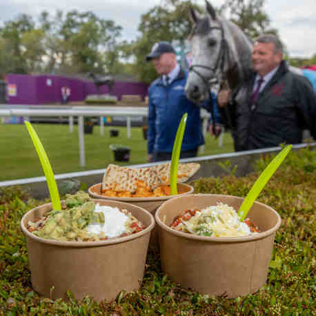 3 bowls of food resting on a hedge whilst 2 men lead a horse in a background at a racecourse.
