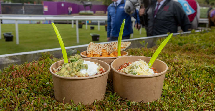 3 bowls of food resting on a hedge whilst 2 men lead a horse in a background at a racecourse.