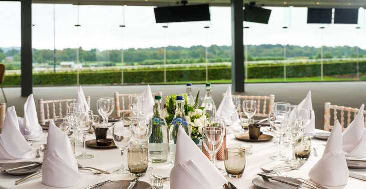 A round table set for dinner with a view of the racecourse through a large window.