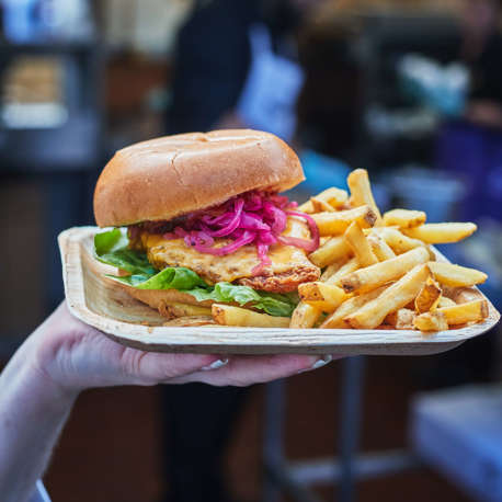 A person's hand holding a cheese burger and fries outside on a small tray.