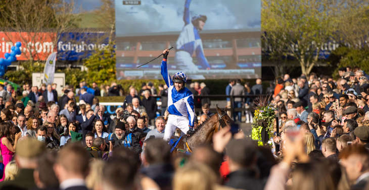 A jockey in a blue and white jersey celebrating winning a race as the crowd looks on and applauds.