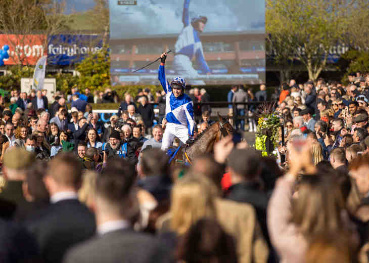 A jockey in a blue and white jersey celebrating winning a race as the crowd looks on and applauds.