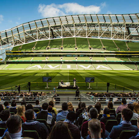 A crowd of people sitting in a stand in an empty Aviva Stadium watching two people speaking at a podium on the pitch.