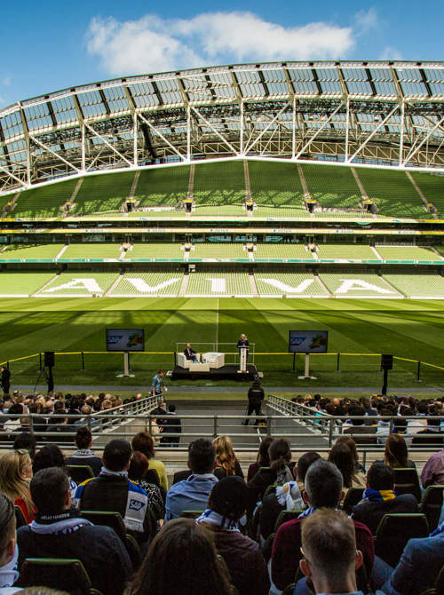 A crowd of people sitting in a stand in an empty Aviva Stadium watching two people speaking at a podium on the pitch.