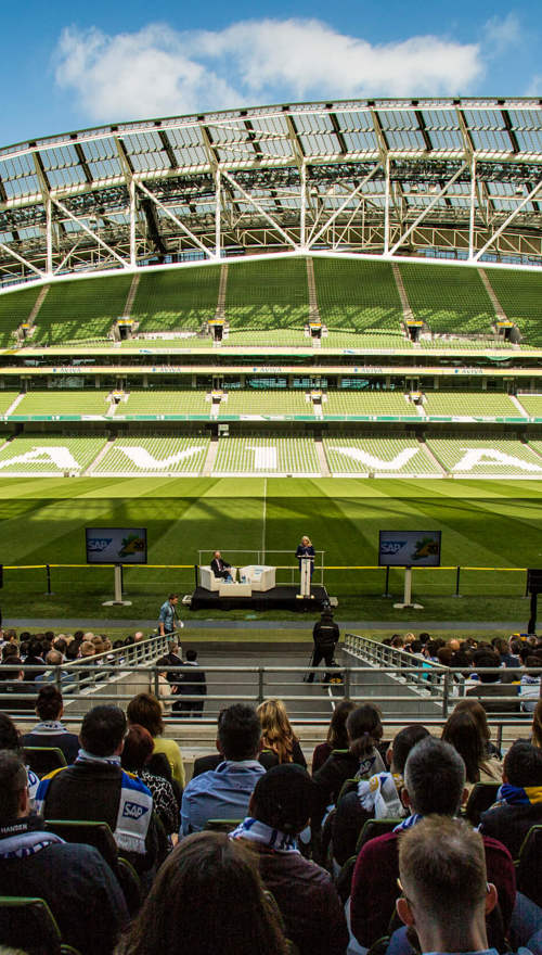 A crowd of people sitting in a stand in an empty Aviva Stadium watching two people speaking at a podium on the pitch.