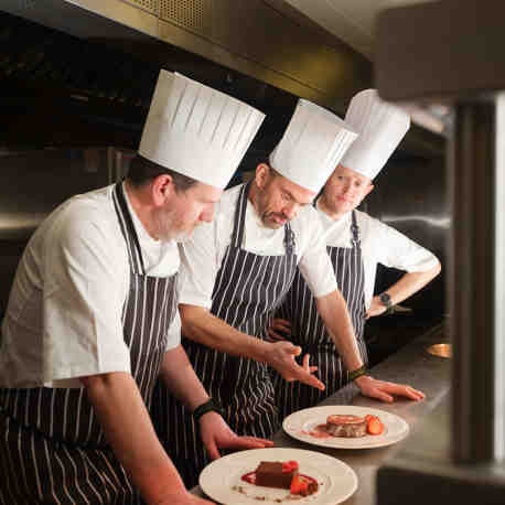 3 chefs talking whilst looking at plates on a kitchen counter.