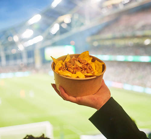 A hand holding a bowl of cheesey nachos with a view of the pitch and a football match at the Aviva Stadium in the background.
