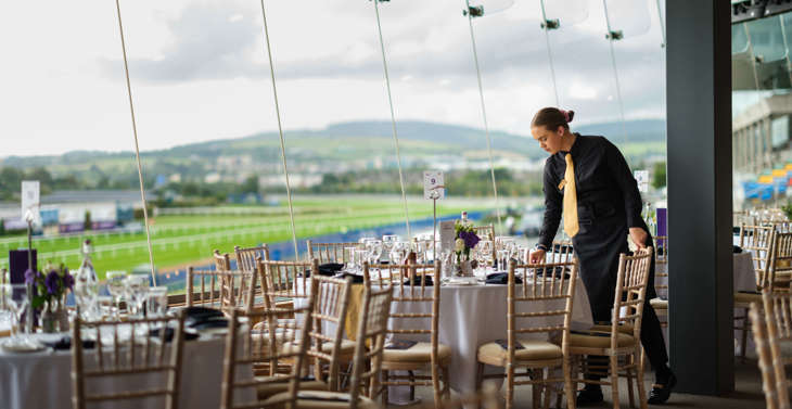 A member of staff setting tables in a room with a view of a racecourse.
