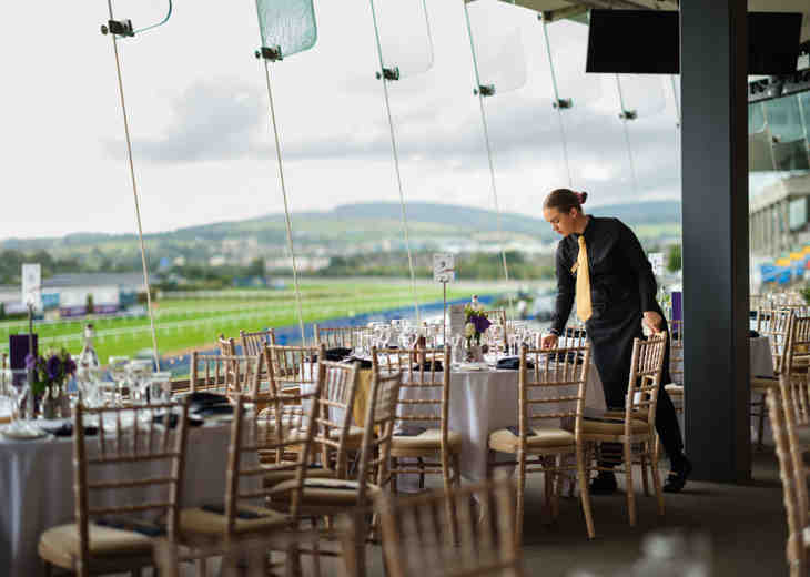 A member of staff setting tables in a room with a view of a racecourse.