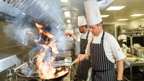 A chef holding a flaming frying pan in a kitchen with another chef behind him.