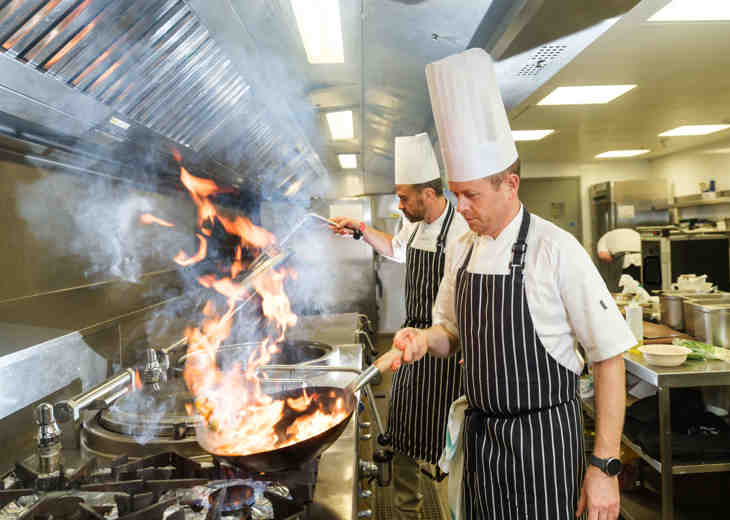 A chef holding a flaming frying pan in a kitchen with another chef behind him.