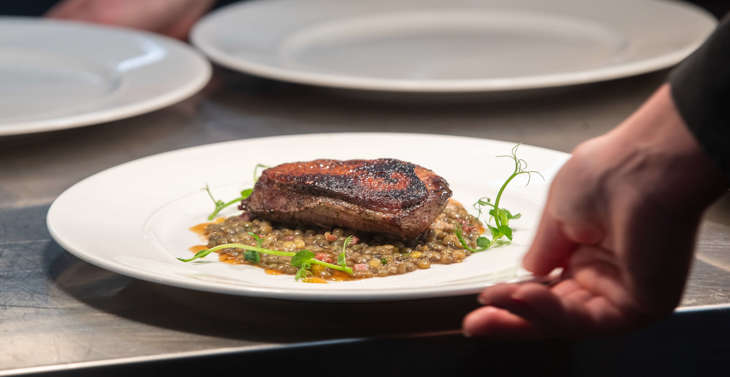A plate of cooked meat served on grains on a metal kitchen counter behind a hand.