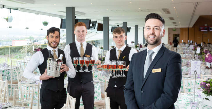 4 male members of staff posing at an event venue with trays of drinks and wine in an ice bucket.