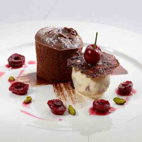 A chocolate cake and a dessert topped with a cherry on a white polate served with cherries.