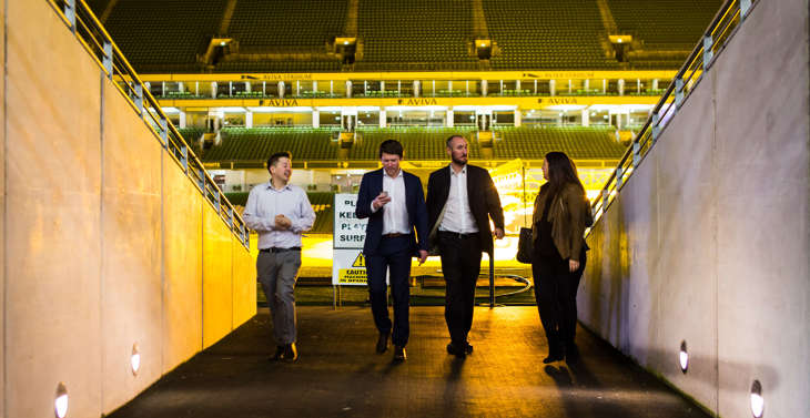 4 people walking down the tunnel at the Aviva Stadium at night with the pitch behind them.