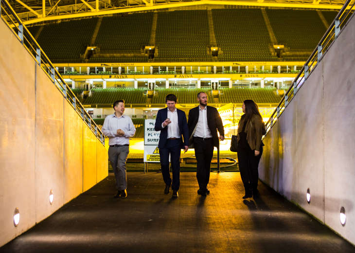 4 people walking down the tunnel at the Aviva Stadium at night with the pitch behind them.