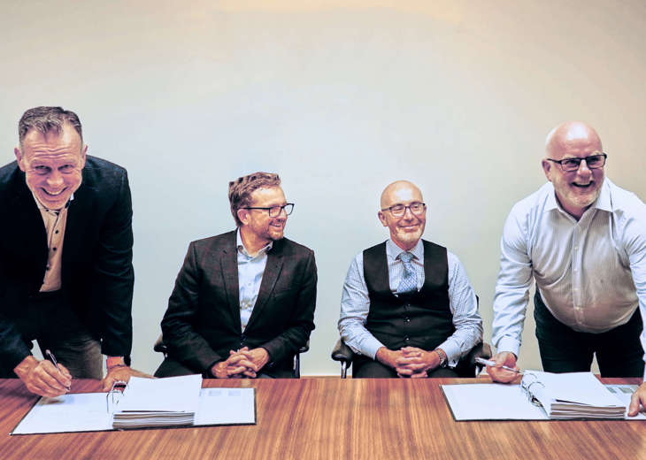 4 men at a table, 2 are seated and 2 are standing signing documents in a ring binder.