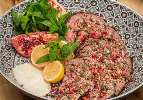 Slices of cooked meat served with lemons, seeds, a sliced pomegranate, leaves and white dip in a bowl on a table.