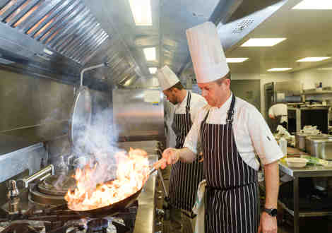 A chef holding a flaming frying pan in a kitchen with another chef stood behind him.