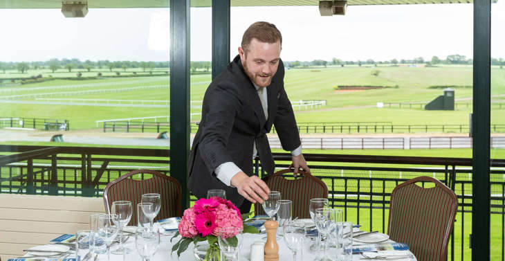 A man arranging glasses on a dining table in a racecourse venue.