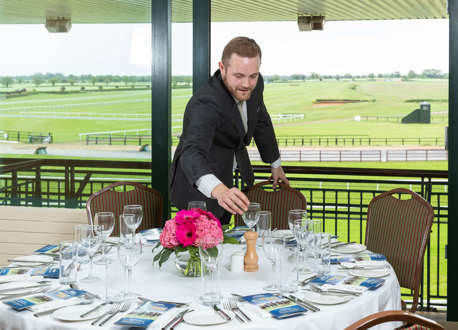 A man arranging glasses on a dining table in a racecourse venue.