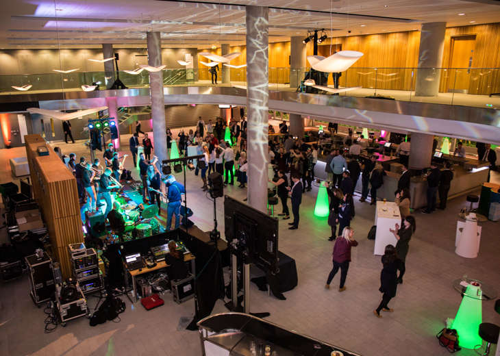 An aerial view of a band playing in a foyer to a crowd of people with people standing at a bar in the background.