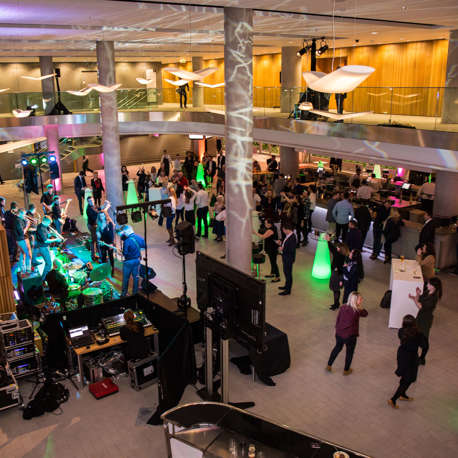 An aerial view of a band playing in a foyer to a crowd of people with people standing at a bar in the background.