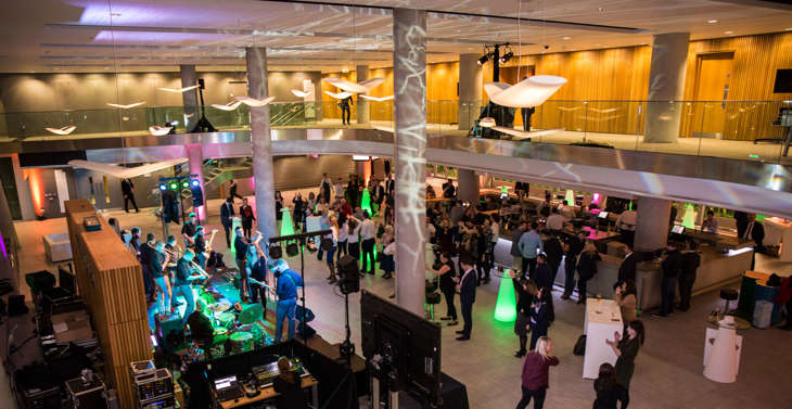 An aerial view of a band playing in a foyer to a crowd of people with people standing at a bar in the background.